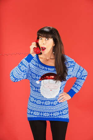Surprised woman in ugly blue Santa Claus sweater with surprised expression on red telephoneの写真素材