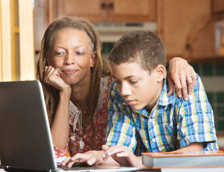 Mother overlooks as her teenaged son uses his laptop to study in their kitchenの写真素材