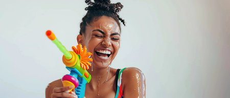 A Laughing young woman in a striped tank top enjoying a moment with a colorful water gun, white backdrop.の素材