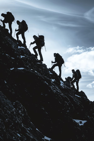 A silhouette of a team of hikers helps each other climb a steep mountain ridge, highlighting themes of teamwork and challengeの素材