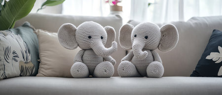 A Two adorable crocheted elephant toys seated on a home sofa with a cozy ambiance, symbolizing handmade crafts and childhood comfortの素材