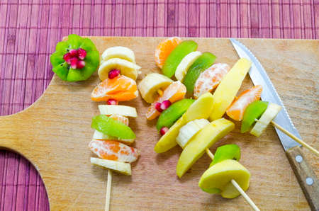Various skewered bananas, oranges and kiwi on a cutting board next to a knifeの写真素材