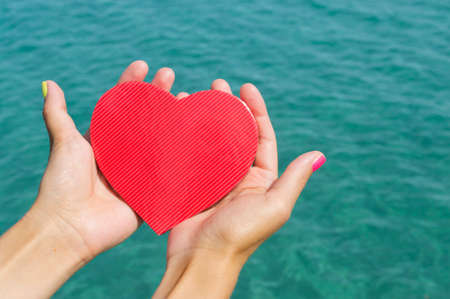 Female hands holding a paper heart above the clear sea waterの写真素材