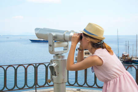 Young girl looking thru public binoculars at the seaside wearing straw hat and pink dressの写真素材