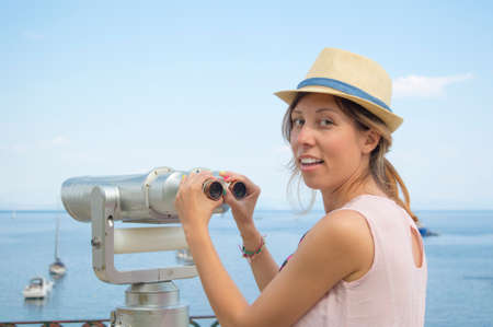 Young girl holding public binoculars at the seaside wearing straw hat and pink dressの写真素材