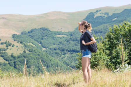 Young, beautiful girl with a backpack on her back, standing on the plateau. Green meadows and majestic mountains in the background. Hiking trip backgroundの写真素材