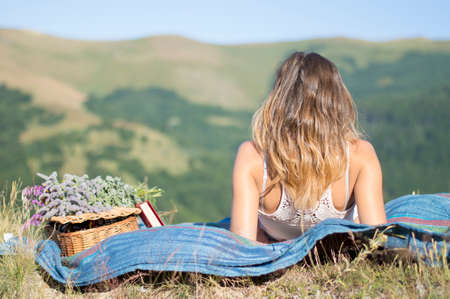 Girl laying on a blanket on a picnic in the field facing the view at the mountainsの写真素材
