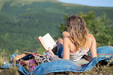 Young woman laying on a blanket and reading a book on a picnic in the field with the view at the mountainの写真素材