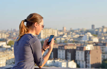 Woman  using her smartphone on rooftop with town panorama in backgroundの写真素材