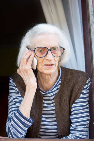 Old grandma talking on the phone while standing on her home windowの写真素材
