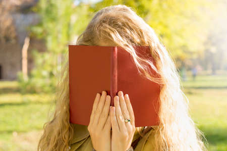Blonde girl covering face with a  red book in a parkの写真素材