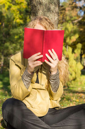 Blonde girl reading a red book in a park on a sunny autumn day