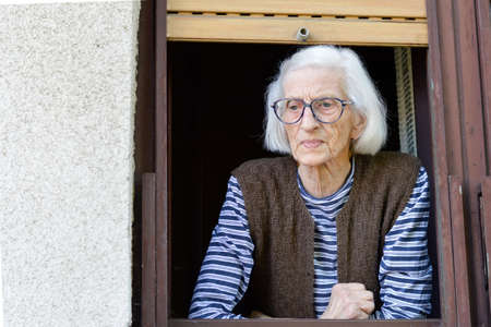 Old and lonely lady looking outside through her window
