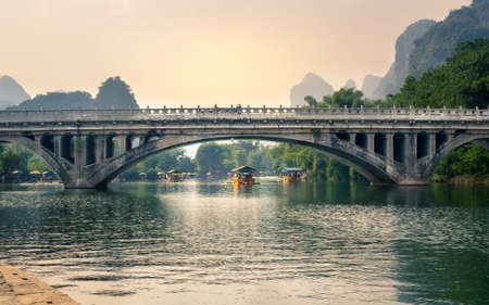 YANGSHUO, CHINA - SEPTEMBER 23, 2016: Boats with tourists floating on Li River, below a stone bridge at sunset timeのeditorial素材