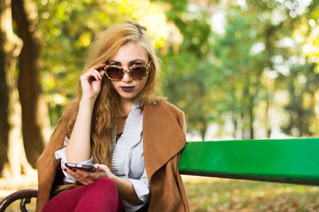 Fashionable girl sitting on a park bench holding a phoneの写真素材