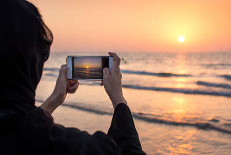 Woman in hijab taking picture of a sunset on the beachの写真素材