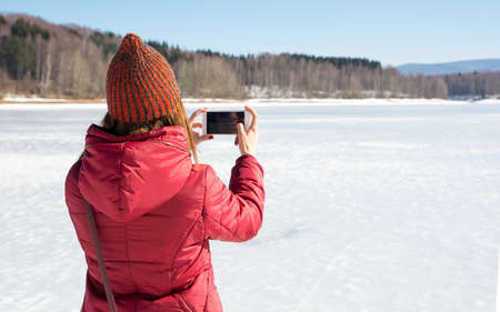 Girl taking picture of a frozen lake with smart phoneの写真素材