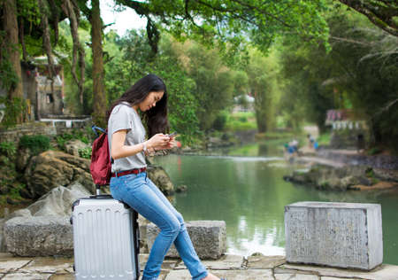 Asian girl siting on the suitcase and playing her phone in the stunning tourism spotの写真素材