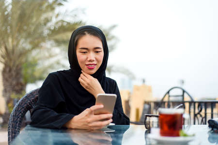 Muslim woman using phone while having a cup of coffee outdoors
