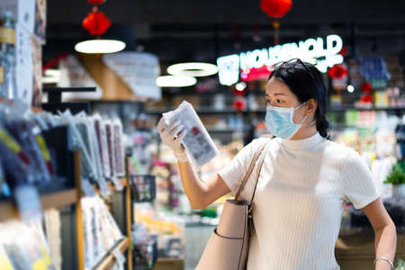 Asian woman shopping for groceries in the market wearing mask and gloves to prevent virius spread during coronavirus pandemicの写真素材