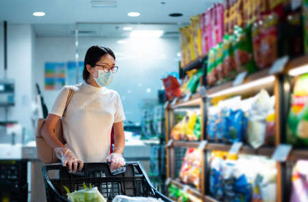 Asian woman shopping for groceries in the market wearing mask and gloves to prevent virius spread during coronavirus pandemicの写真素材