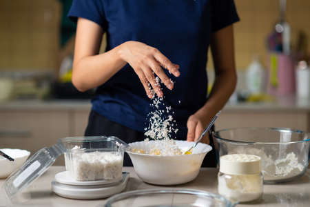 Woman adding grated coconut into cookie dough while making homemade cookies in the kitchen at home...