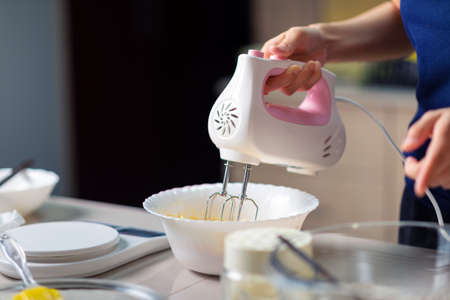 Woman using electric mixer to mix ingredients for dough of sugar, beaten eggs, butter and flour...
