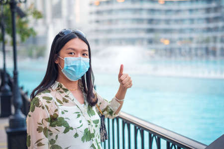Happy female Asian tourist wearing face mask and taking photo at Dubai mall fountain outdoors. Visiting shopping mall, travel and leisure new normal after coronavirus Covid-19 pandemic lifestyleの写真素材