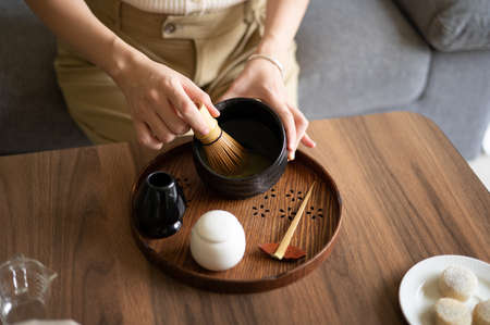 Woman mixing Japanese matcha green tea beverage at home using traditional tea ceremony bamboo set and natural whisk while sitting on the sofa at home.の写真素材