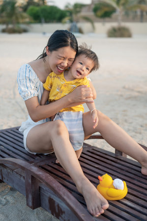 Baby boy on a beach holiday sitting on the sunbed with his mother at sunset. Asian woman and her one...