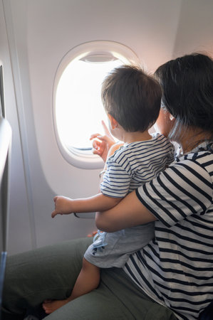 Infant traveling in airplane sitting on its mother lap both looking out of an airplane window. One...