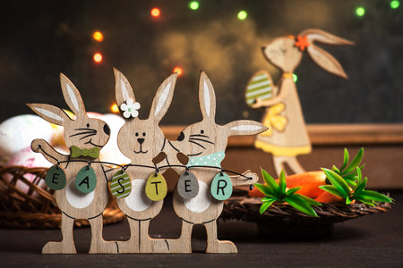 Three wooden Easter bunnies holding Easter eggs displayed as decorations on a wooden table in front of a window with festive decorationsの写真素材
