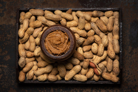 Creamy, freshly made peanut butter in a wooden jar and heaps of shelled peanuts around