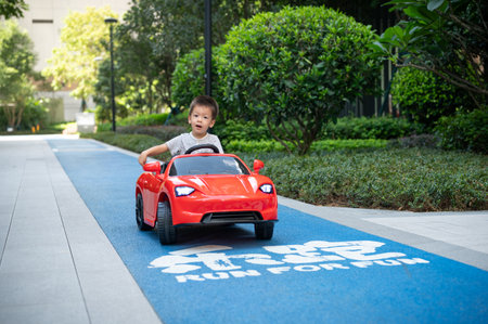 Cute multiracial toddler boy looks around while riding a red remote controlled car on a paved trackの写真素材