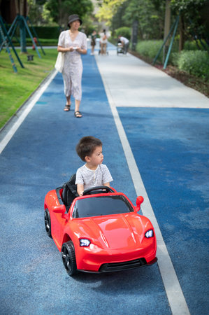 Cute multiracial toddler boy rides in a red remote controlled car while his mother walks behind him driving a toy vehicleの写真素材