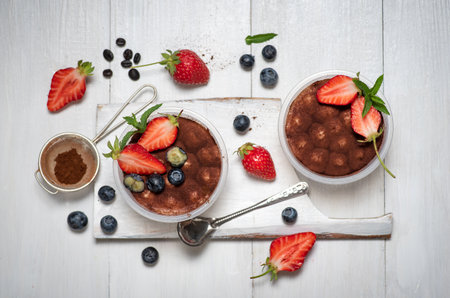 Traditional Italian sweet dessert of tiramisu cake in glass on white wooden table. With cocoa powder and a refreshing addition of strawberry and blueberryの写真素材