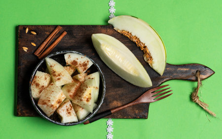 Slices and small pieces of ivory gay melon sprinkled with cinnamon in a bowl, on a cutting board and a green background.Ivory gaya melon with green dotted stripes and spots. Colorful ripe juicy and soft fruit, sweet taste with floral notesの写真素材