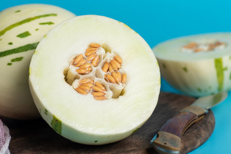 Ivory gaya melon with green dotted stripes and spots on a blue background. Colorful ripe juicy and soft fruit, sweet taste with floral notes. Whole and half melons on a wooden cutting board, close upの写真素材
