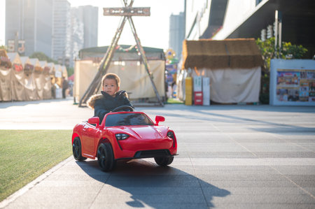 A young multiracial toddler joyfully navigates the neighborhood road in a red sports toy car,...