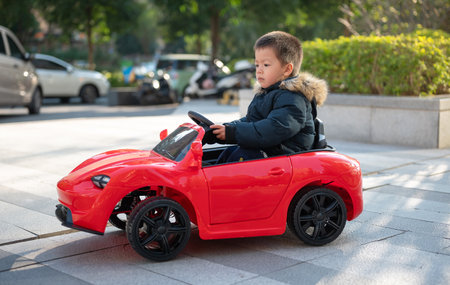 A young multiracial toddler joyfully navigates the neighborhood road in a red sports toy car,...