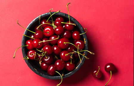 A striking image featuring a black bowl filled with fresh, ripe cherries set against a stark red background. The vivid contrast between the dark bowl and the bright cherries enhances the image's visual appeal, making it an ideal representation of freshness and simplicity, perfect for food-related themes.の写真素材