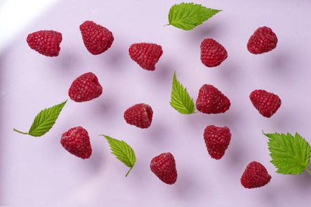 Juicy raspberries and bright green leaves are artfully arranged on a pink background, creating a...
