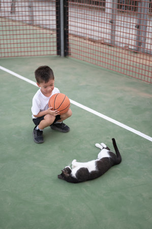 4 year old boy is kneeling on a basketball court, holding a basketball, while a playful cat lies on the ground nearby, capturing a charming moment of childhood joy.の写真素材