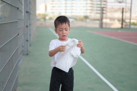 A focused young boy stands on a basketball court, wiping his brow with a towel after a game, conveying determination and the spirit of sportsmanship.の写真素材