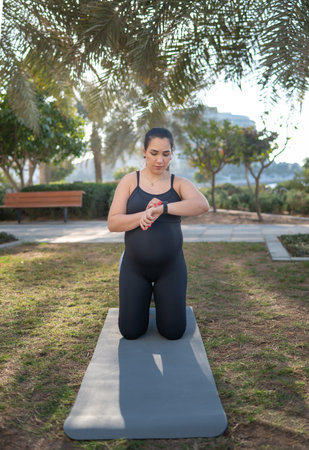 A pregnant woman in black athletic wear kneels on a yoga mat in a sunlit park, checking her watch. The scene conveys fitness, maternity wellness, and calm focus during outdoor exercise.の写真素材