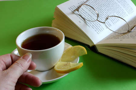 Hand reaching for cup of tea with lemon and book with glassesの写真素材