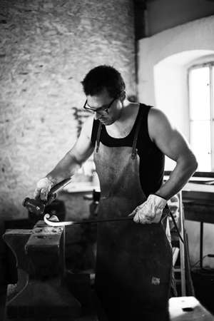 The blacksmith in the production process of metal products handmade in the forge. Blacksmith forging metal with a hammer. Black and white photoの写真素材