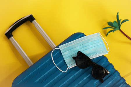 Travel during the time of COVID-19. Medical mask, suitcase, sun glasses, seashells on the yellow background. Vacation, holidays in corona times.の写真素材
