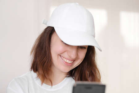 Female model wearing a white baseball cap. White cap mockup, template for picture, text. Girl with a cap holding smarthphone. Free space, copy space.の写真素材
