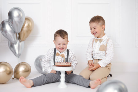 Two boys celebrating birthday, children have a B-day party. Birthday cake with candles and balloons. Happy kids, celebration, white minimalist interior.の写真素材